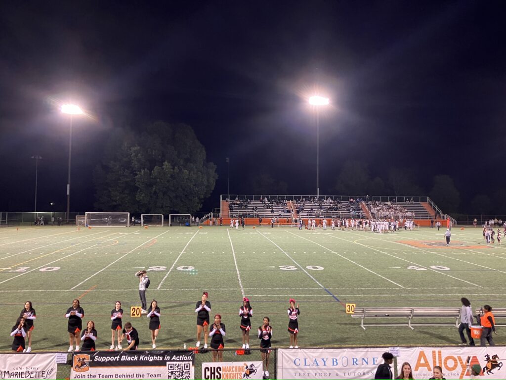 A group of cheerleaders perform for an audience on a football field at night.