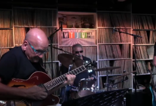 A group of five jazz musicians play music in a dimly lit space with shelves of vinyl records behind them.