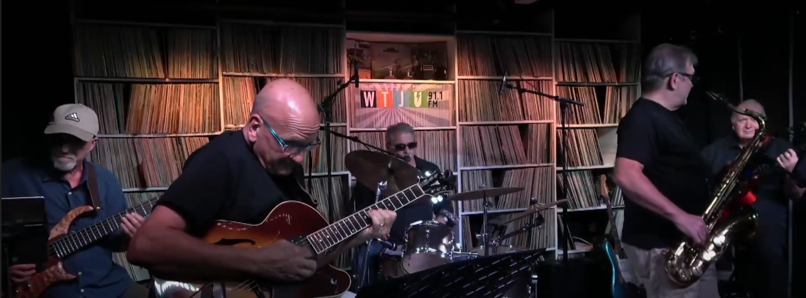 A group of five jazz musicians play music in a dimly lit space with shelves of vinyl records behind them.