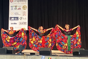 Three dancers stand on a stage wearing ornate dresses.