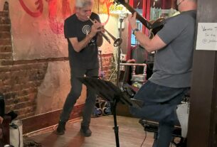 A gray-haired man in black clothing playing trumpet facing another man playing guitar