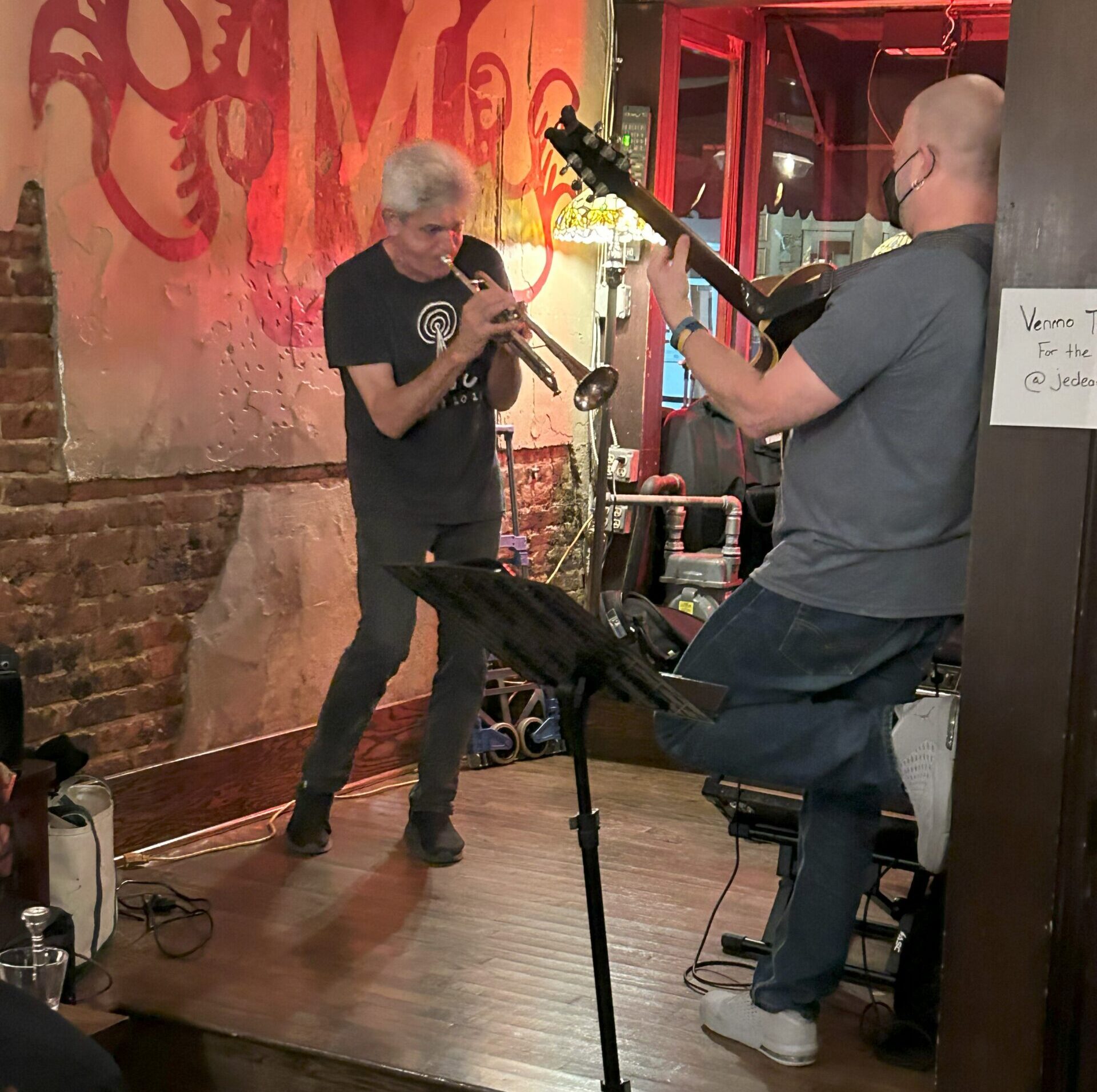 A gray-haired man in black clothing playing trumpet facing another man playing guitar