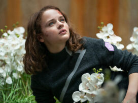 A teenage girl with bruises and scabs on her face lays in a patch of white flowers.