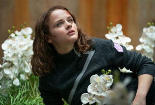 A teenage girl with bruises and scabs on her face lays in a patch of white flowers.