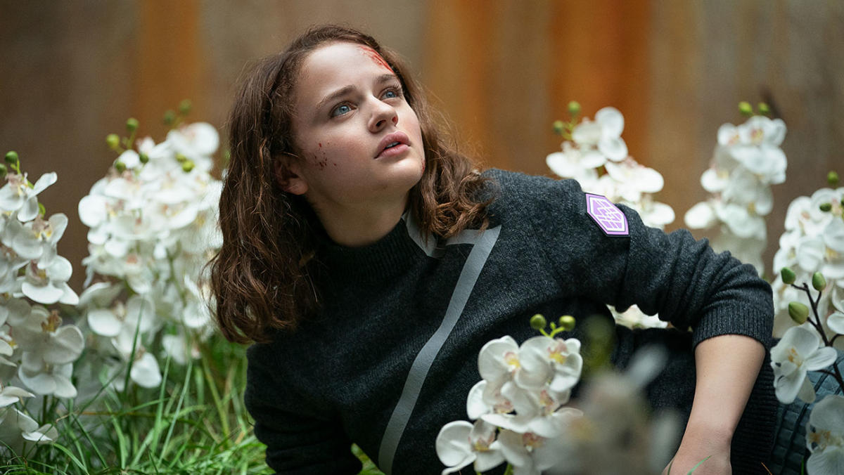 A teenage girl with bruises and scabs on her face lays in a patch of white flowers.