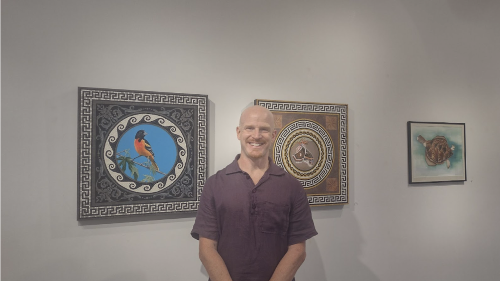 A man in a purple polo shirt stands in front of his art.