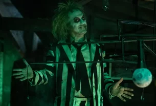 Beetlejuice, played by Michael Keaton stands with his arms outstretched in dim green light.