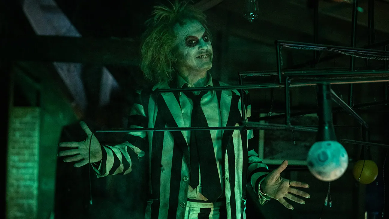 Beetlejuice, played by Michael Keaton stands with his arms outstretched in dim green light.