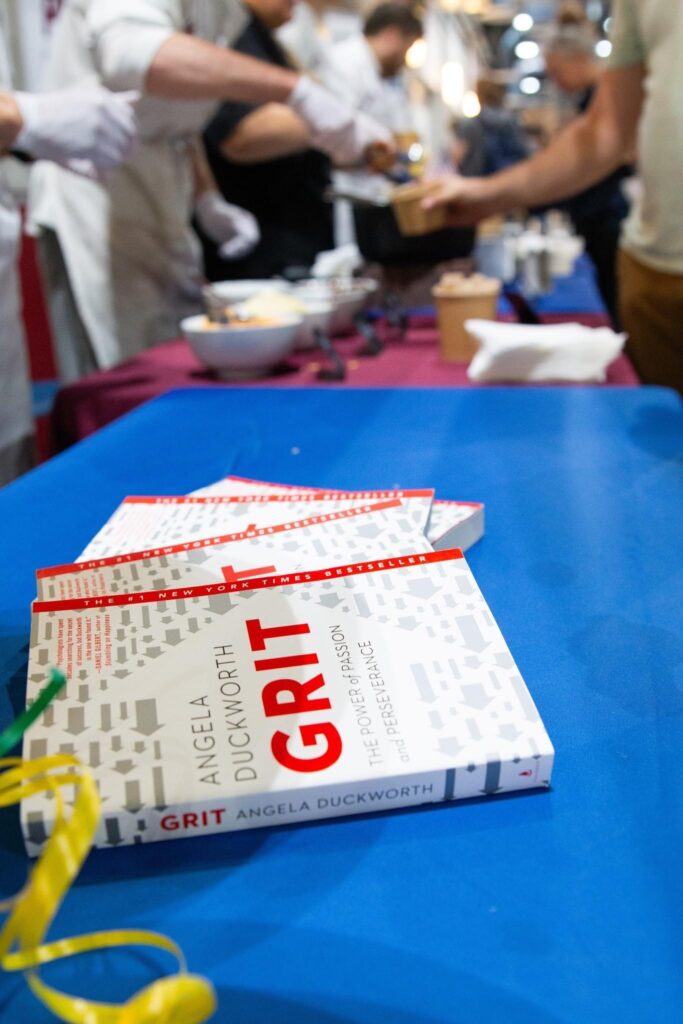 Several copies of Angela Duckworth's "Grit: The Power of Passion and Perseverance" sit on a table where Culinary Arts students feed people grits.