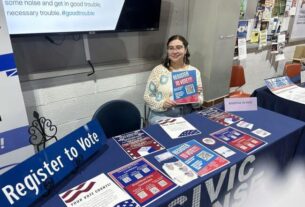 A female student sits at a table with a blue tablecloth holding a paper that says register to vote. The table has a civic sense logo on it