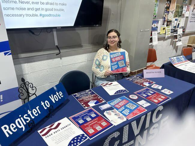 A female student sits at a table with a blue tablecloth holding a paper that says register to vote. The table has a civic sense logo on it