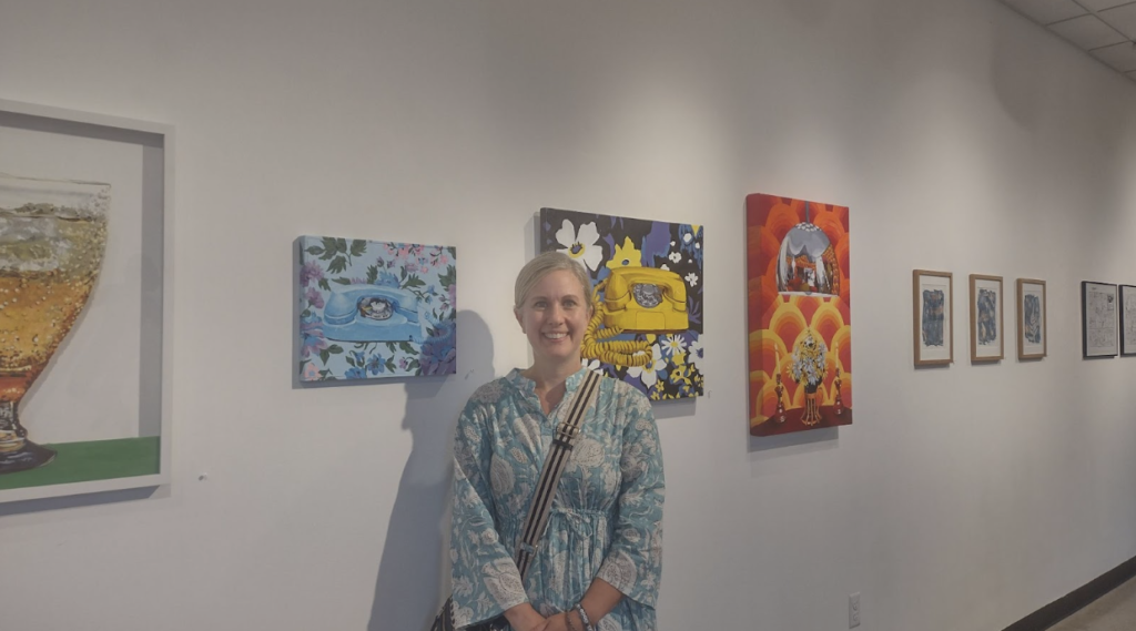 A woman in a floral dress stands in front of 3 of her art pieces.