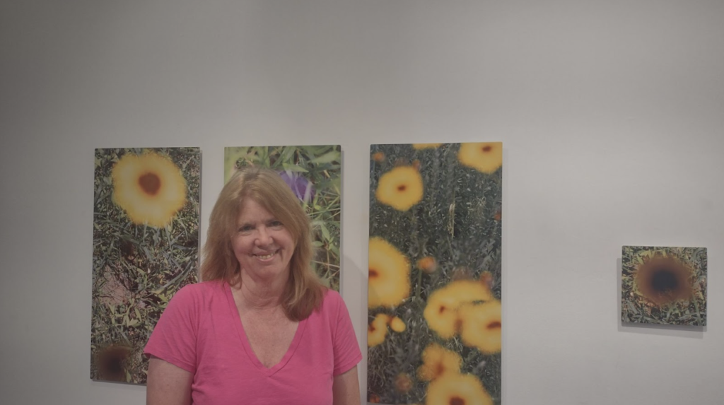 A woman wearing a pink V-neck stands in front of her artwork, which are photos of yellow flowers.