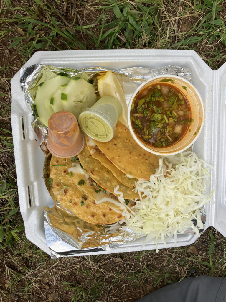 A takeout box with four street tacos, cucumbers and consumé.