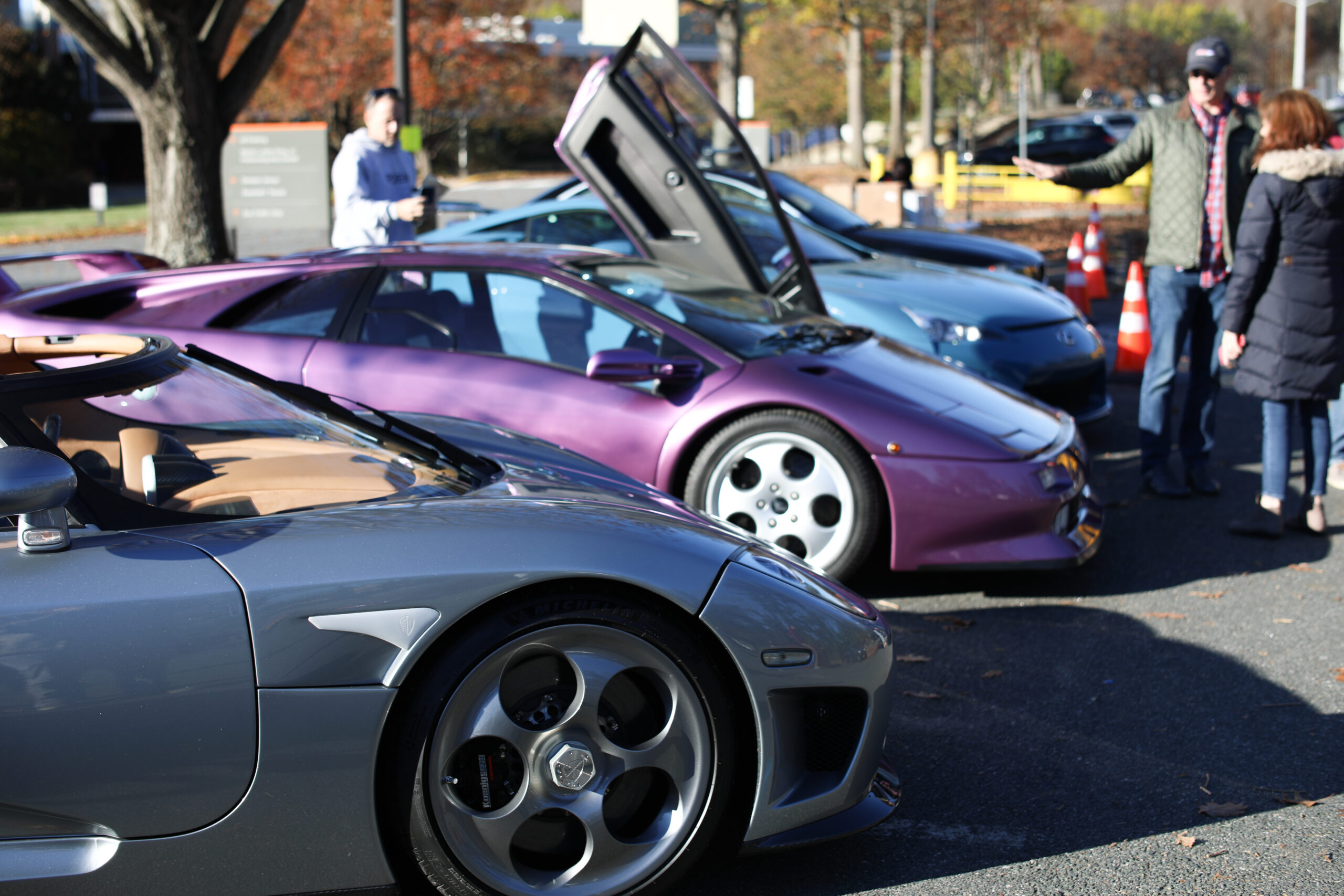 Three supercars are parked side by side on main display in a parking lot. Left to Right: Grey Koenigsegg CCXR, Purple Lamborghini Diablo SE30, Powder Blue Lexus LFA.