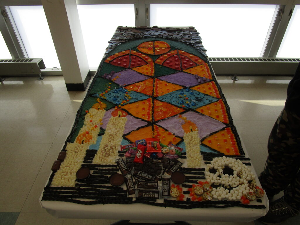 A mosaic made of candy showcases candles alight and a skull in front of a set of a colored window tiles.