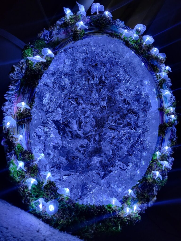 A blue oval of swirled glass lit in light blue surrounded by moss and clear mushroom fairy lights in the darkness.