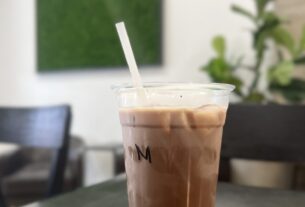 An iced coffee sits on a wood table in the foreground with a green plant wall and armchairs in the background.