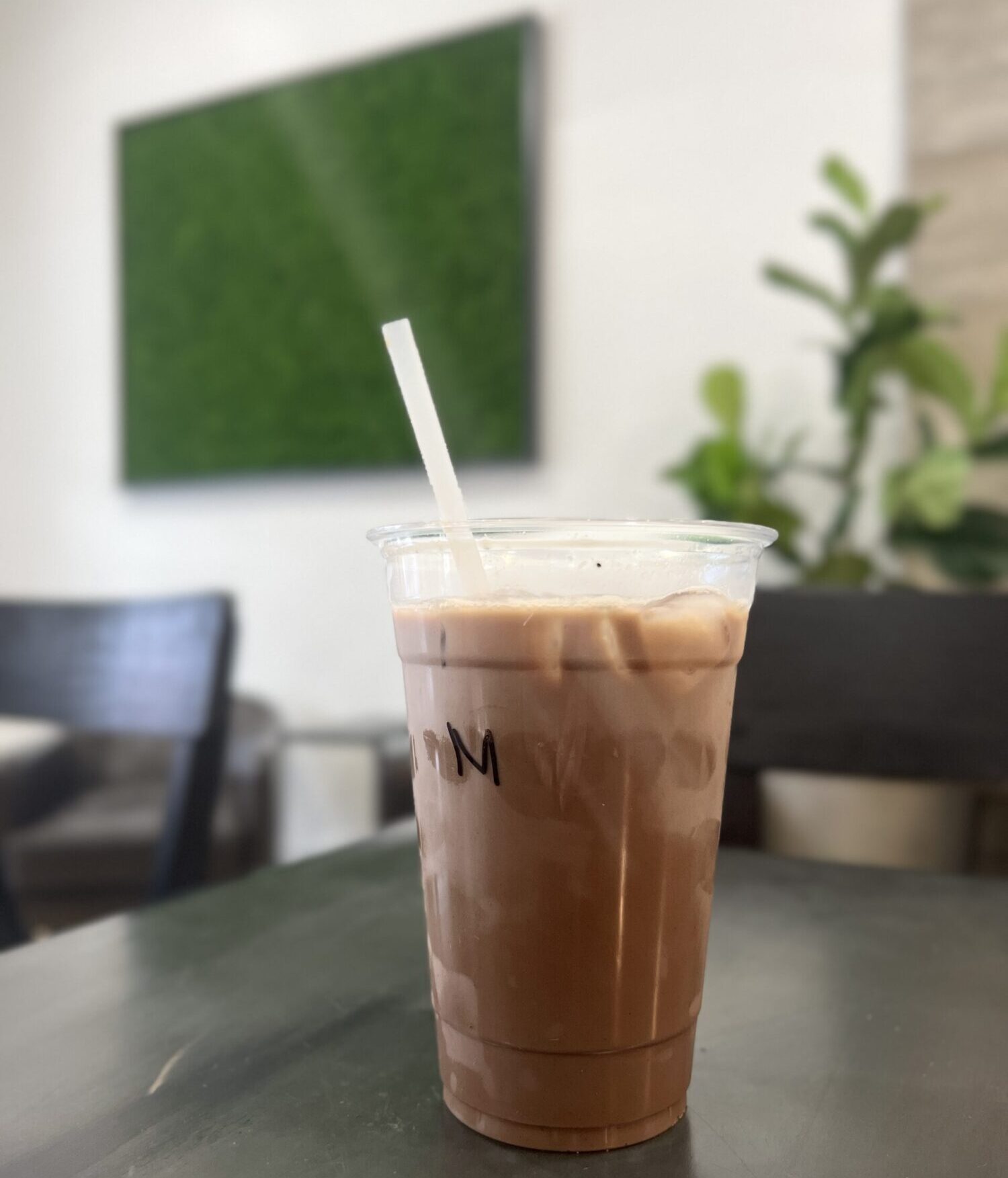 An iced coffee sits on a wood table in the foreground with a green plant wall and armchairs in the background.