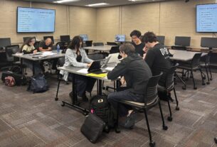 Students sit at desks with their laptops open. TV screens on the wall showcase which articles need to be edited.