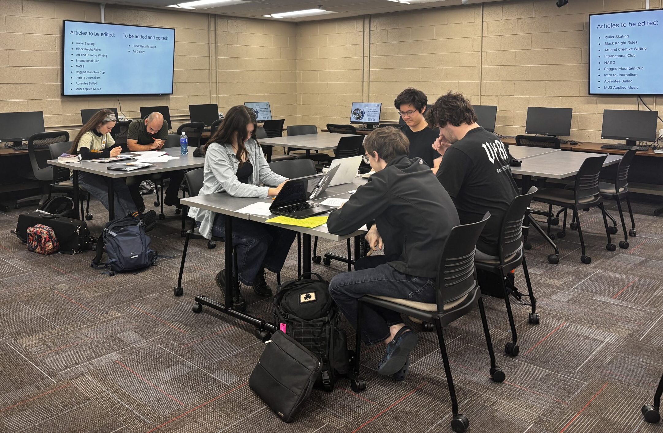 Students sit at desks with their laptops open. TV screens on the wall showcase which articles need to be edited.