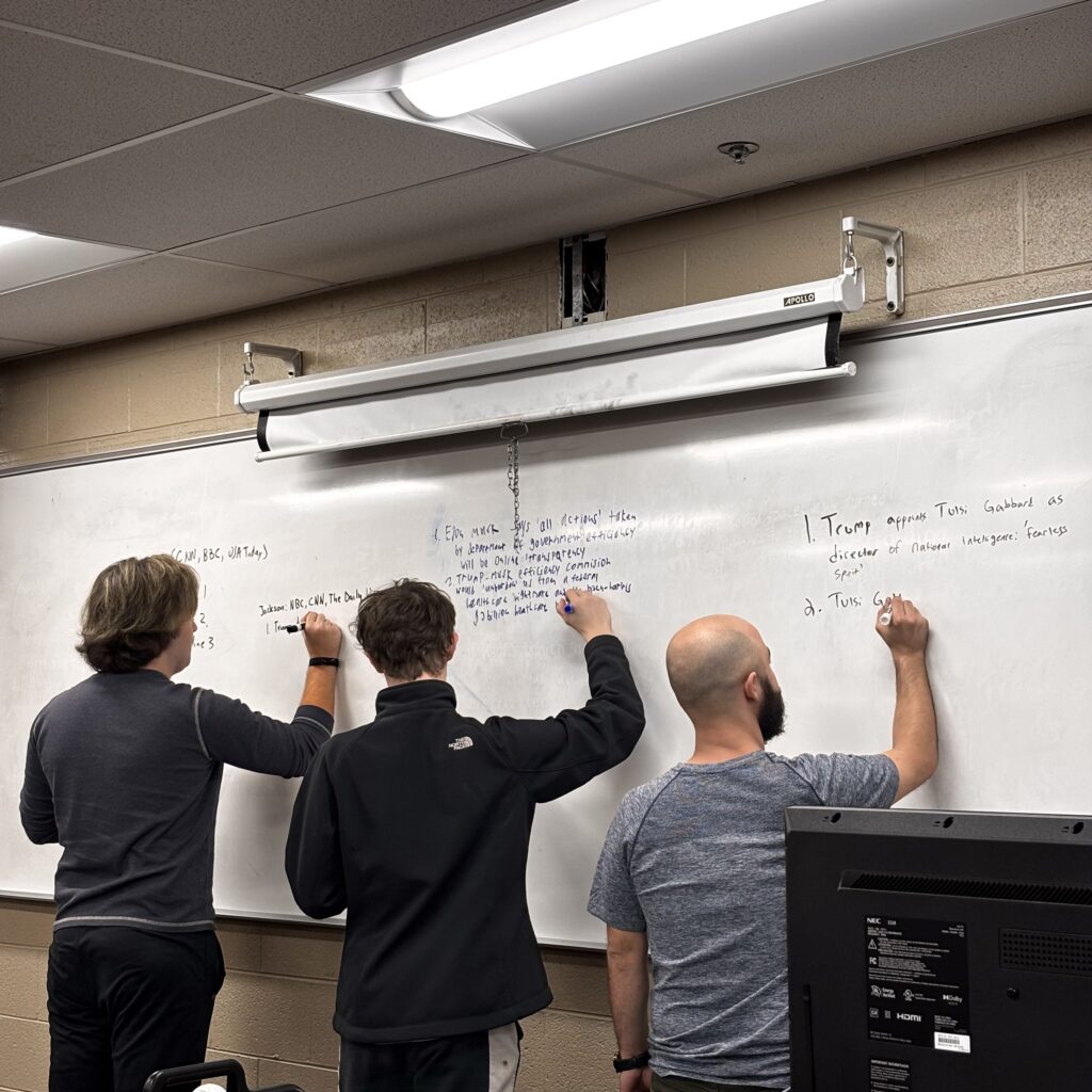 Three students write headlines from media sources on a whiteboard.