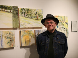 A man in a denim jacket and fedora with his various watercolor and ink sketch works.