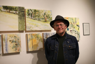 A man in a denim jacket and fedora with his various watercolor and ink sketch works.