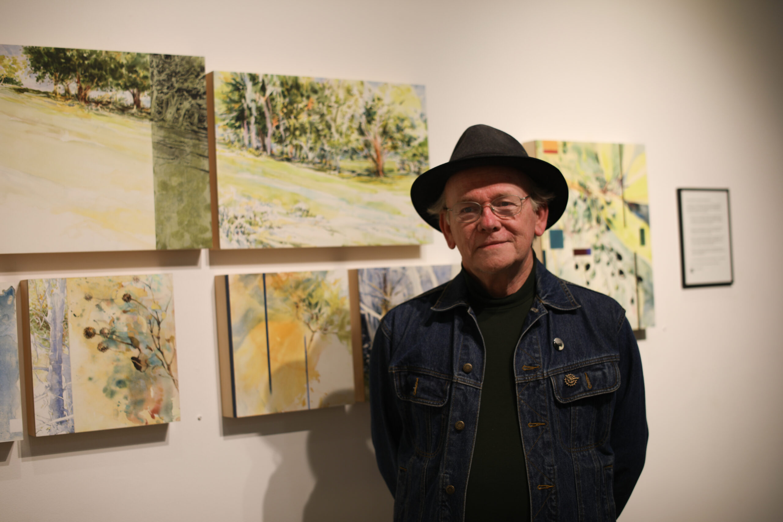 A man in a denim jacket and fedora with his various watercolor and ink sketch works.