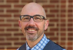 A headshot of a bald white man with black glasses smiling at the camera