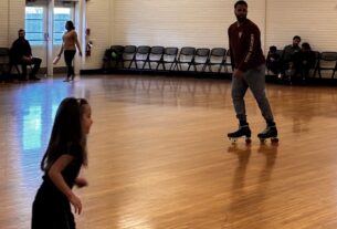 A young girl and a man ride on roller skates on a wooden floor in a white brick building.