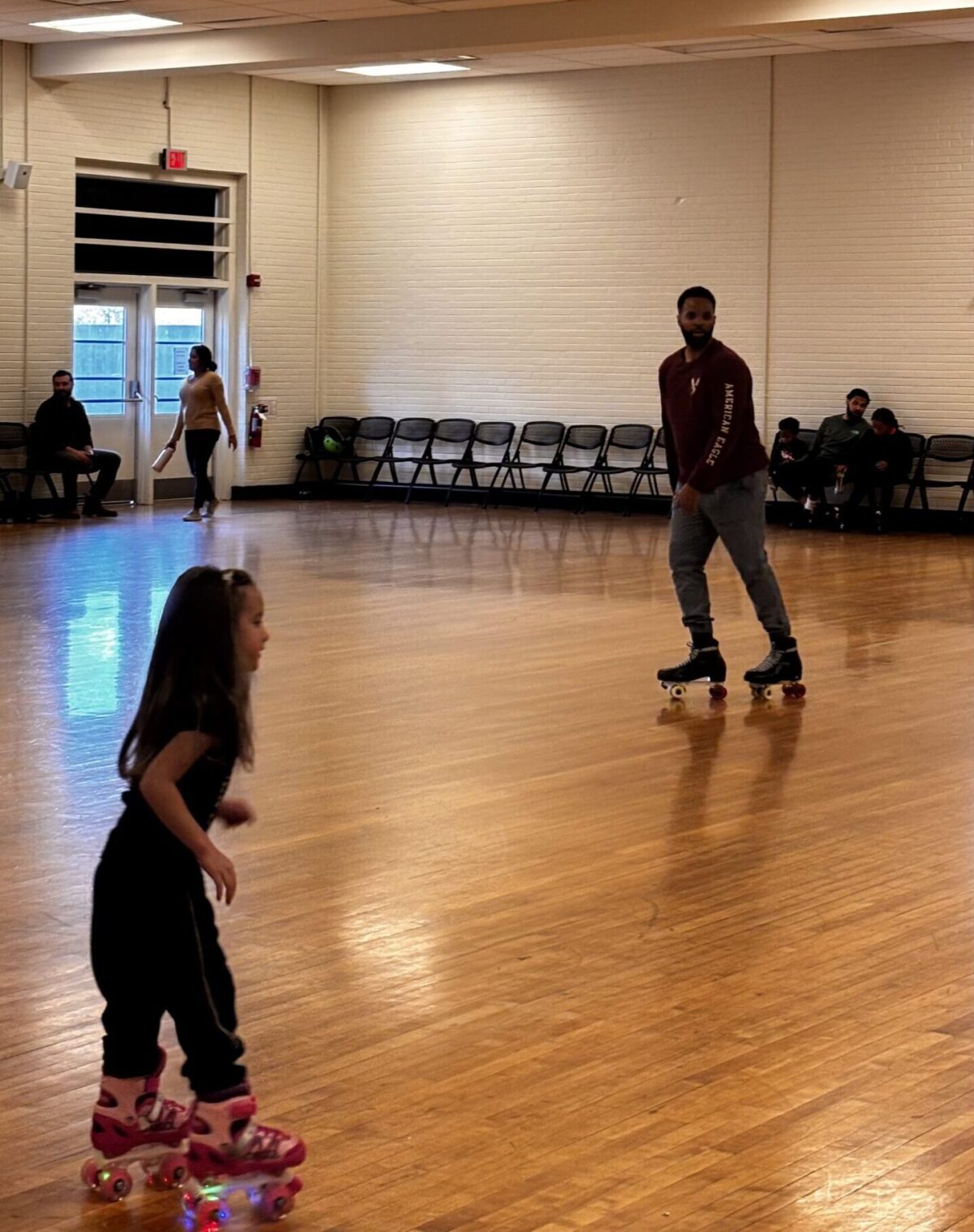 A young girl and a man ride on roller skates on a wooden floor in a white brick building.