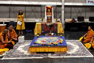 Mary-Evelyn Sellars stands behind the four monks sitting around the sand mandala before the event begins.