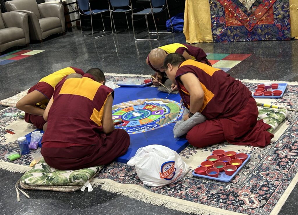 Tibetan monks work on their sand mandala on an ornate rug with cushions to sit on.