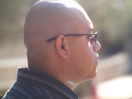 Side prole shot of a bald young man with glasses wearing a black collared shirt on a sunny day.