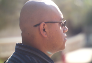 Side prole shot of a bald young man with glasses wearing a black collared shirt on a sunny day.