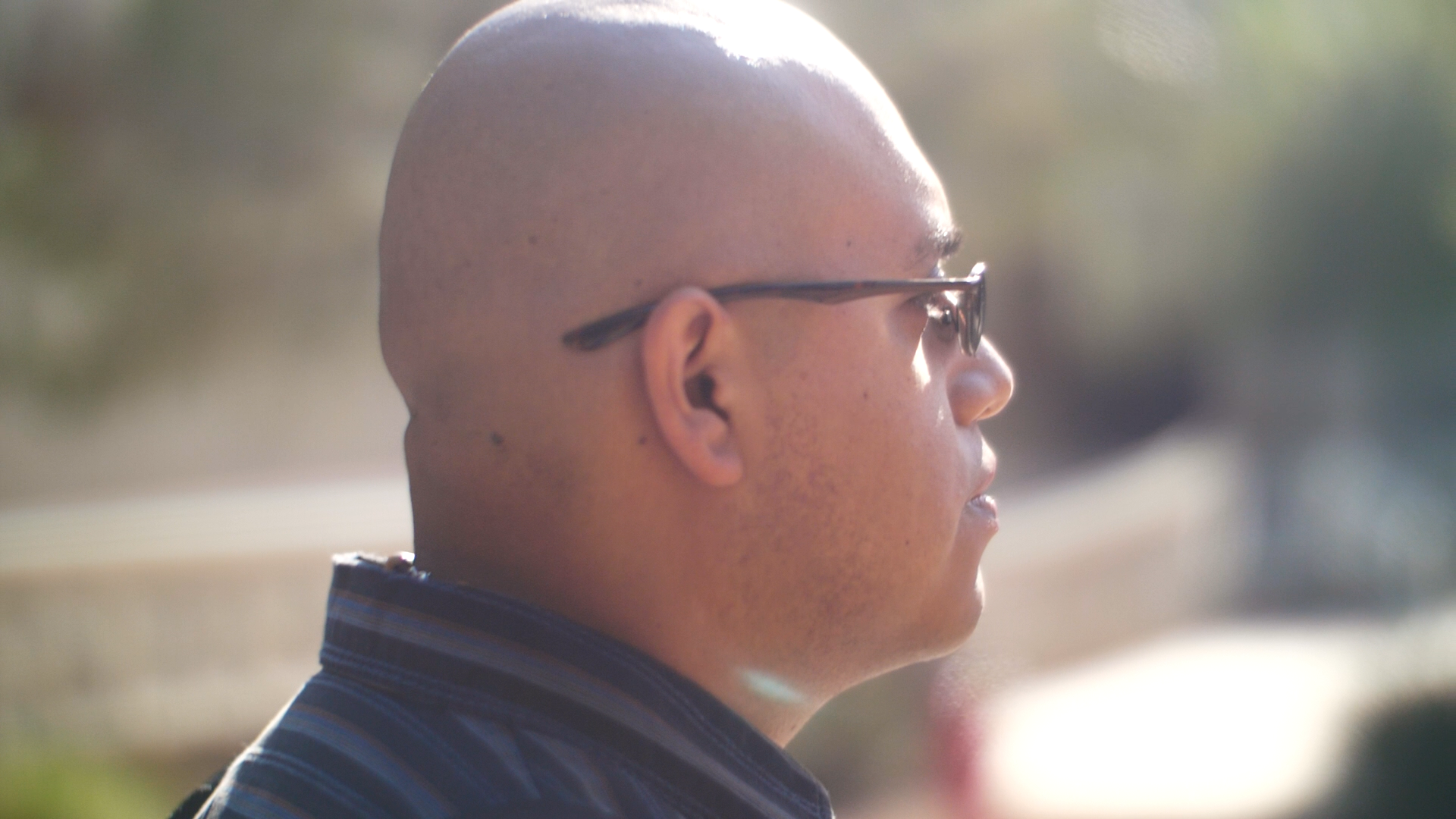 Side prole shot of a bald young man with glasses wearing a black collared shirt on a sunny day.